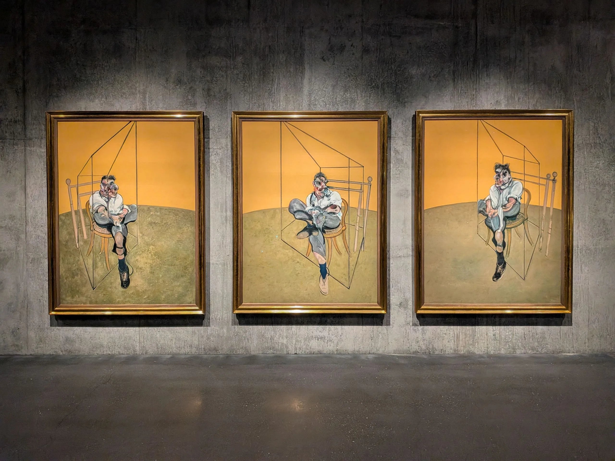Three connected canvases, each showing a contorted, smeared portrait of the painter Lucian Freud seated on a bentwood chair against flat, saturated colour. The distortion is characteristic of Bacon's later portraits of the friends and rivals who sat for him.