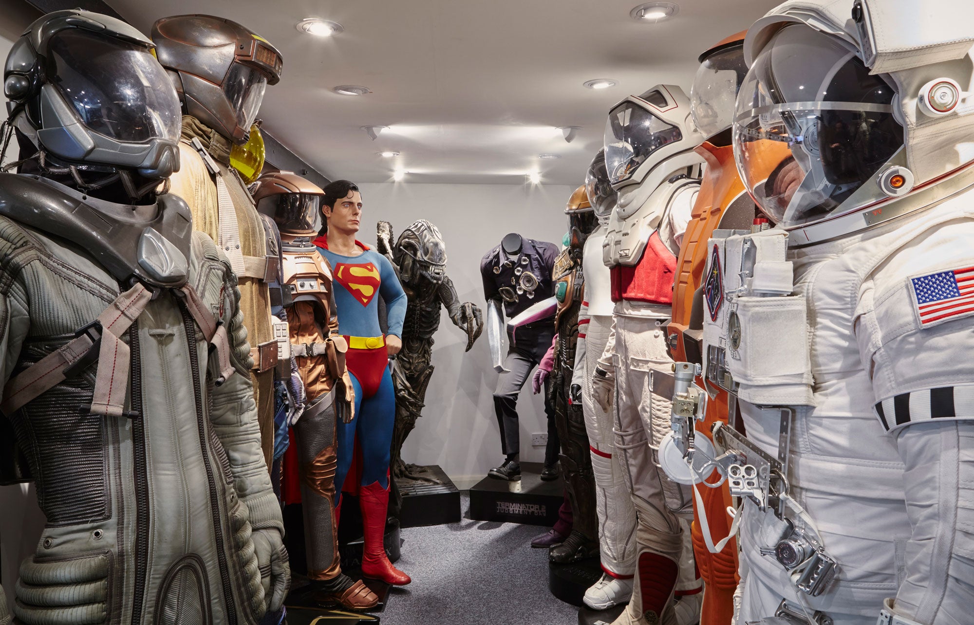 Iconic film costumes on display, featuring various spacesuits, a Superman outfit, a Xenomorph model, and a damaged police uniform from Terminator 2.